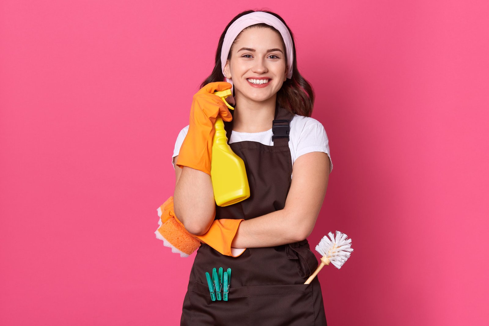 Home housekeeper woman holding bottle with cleaner liquid in hands, female wearing white t shirt and brow apron, looking smiling directly at camera, posing against pink wall.
